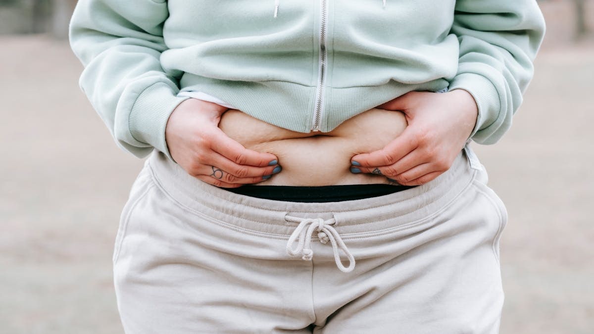 Crop anonymous plus sized female in casual hoodie touching and showing fat on belly while standing in park